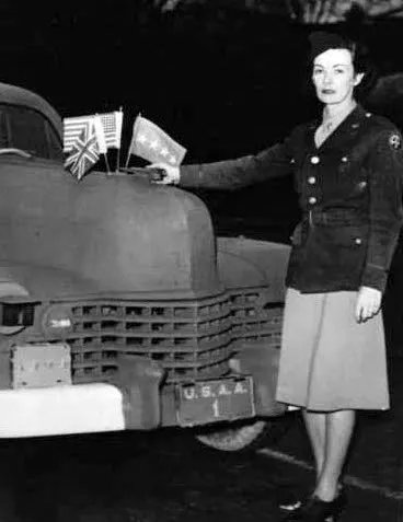 Woman in uniform leaning on a car that has flags on the front hood.